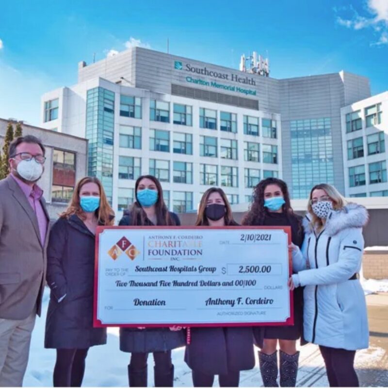 Group of six people holding a large donation check outside Charlton Memorial Hospital. Snow on the ground.
