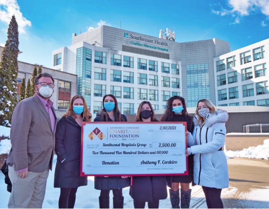 Group of six people holding a large donation check outside Charlton Memorial Hospital. Snow on the ground.