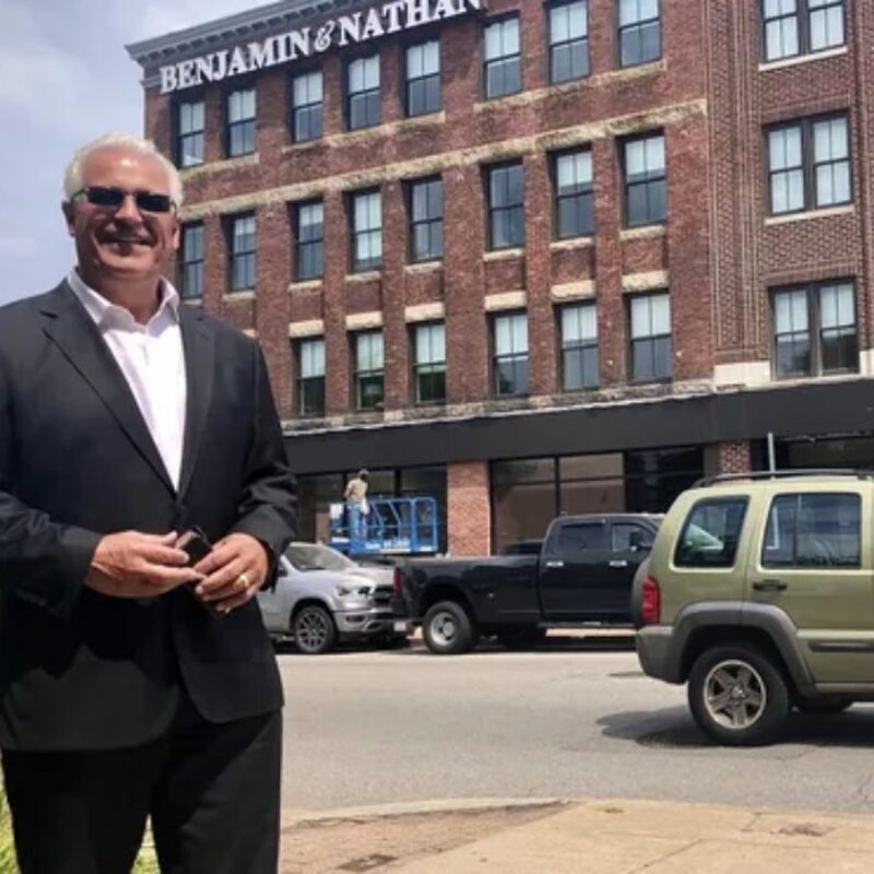 A man in a suit stands in front of a brick building, smiling, with vehicles and a worker on a lift nearby.