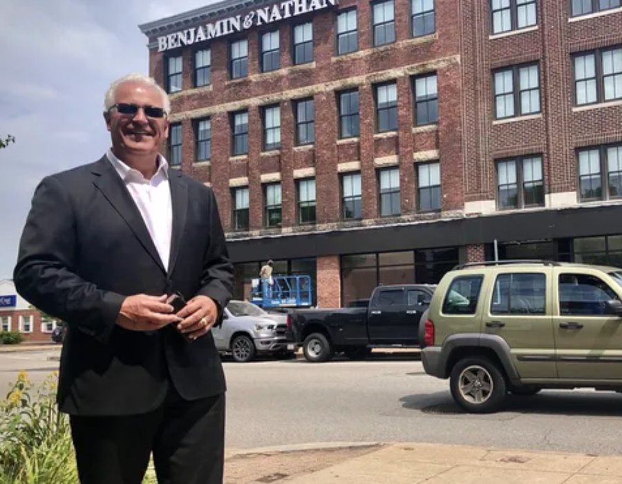 A man in a suit stands in front of a brick building, smiling, with vehicles and a worker on a lift nearby.