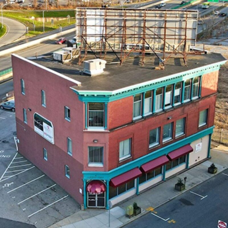 Three-story brick building with teal trim, red awnings, and a large billboard on the roof. Nearby parking area.