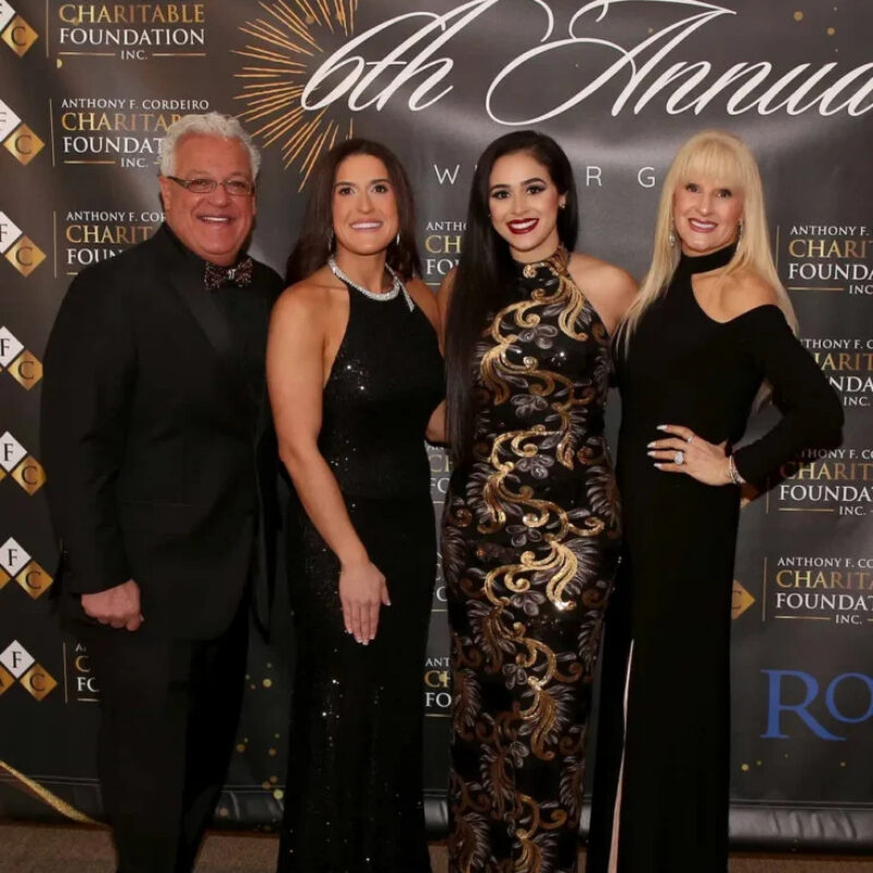 Four elegantly dressed individuals pose together in front of a decorative backdrop for a charity event.