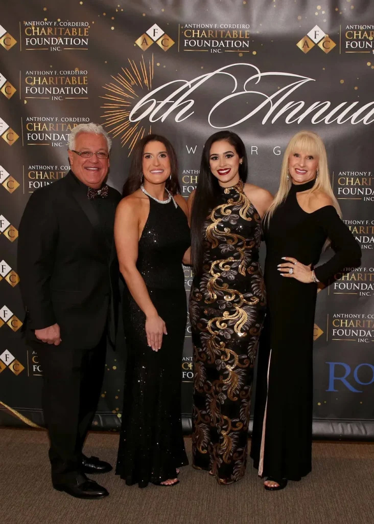 Four elegantly dressed individuals pose together in front of a decorative backdrop for a charity event.