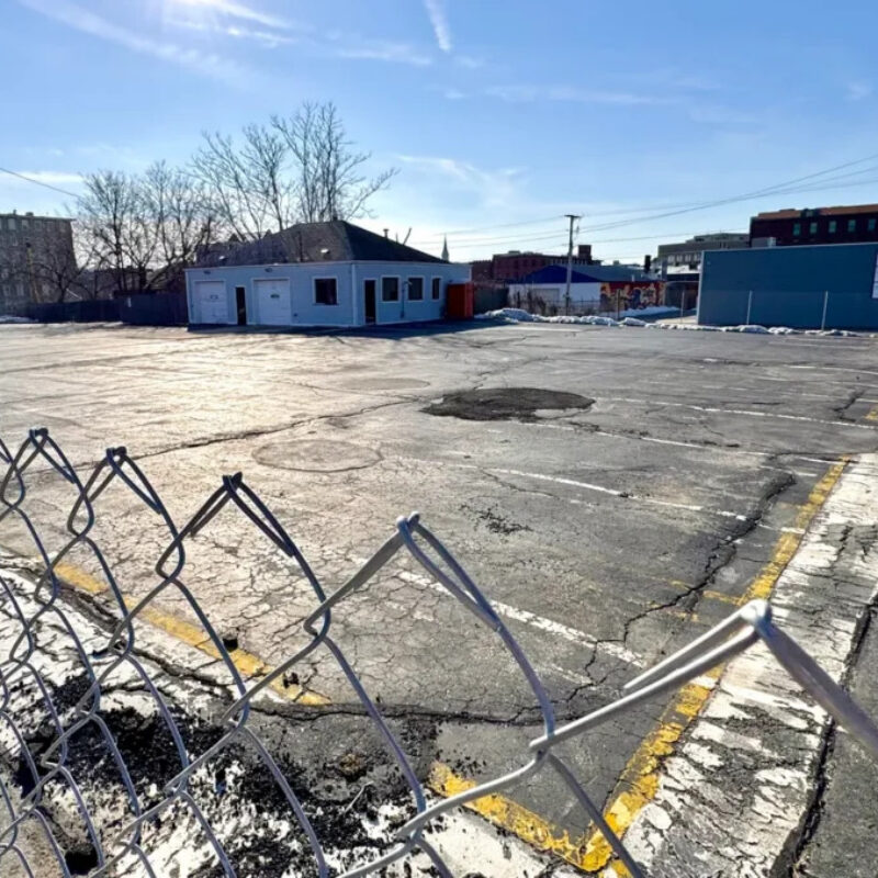 Empty parking lot with a small building and a chain-link fence under a clear blue sky.