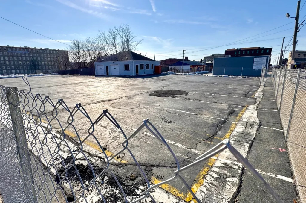 Empty parking lot with a small building and a chain-link fence under a clear blue sky.