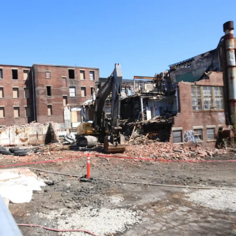 A construction vehicle demolishes a partially collapsed brick building surrounded by debris and safety barriers.