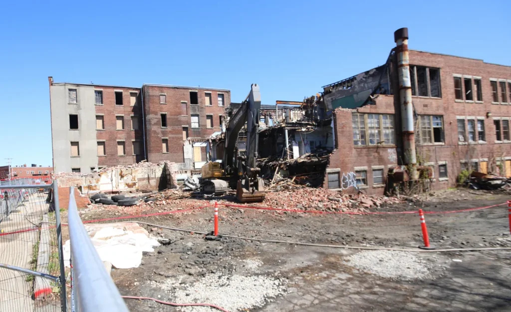 A construction vehicle demolishes a partially collapsed brick building surrounded by debris and safety barriers.