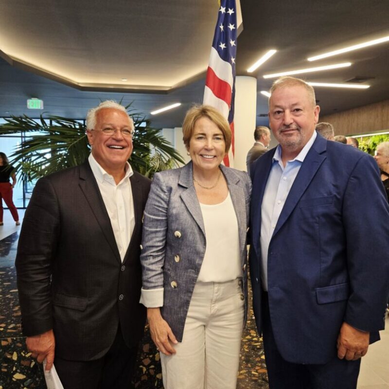 Three people pose together in a modern setting, with an American flag in the background.