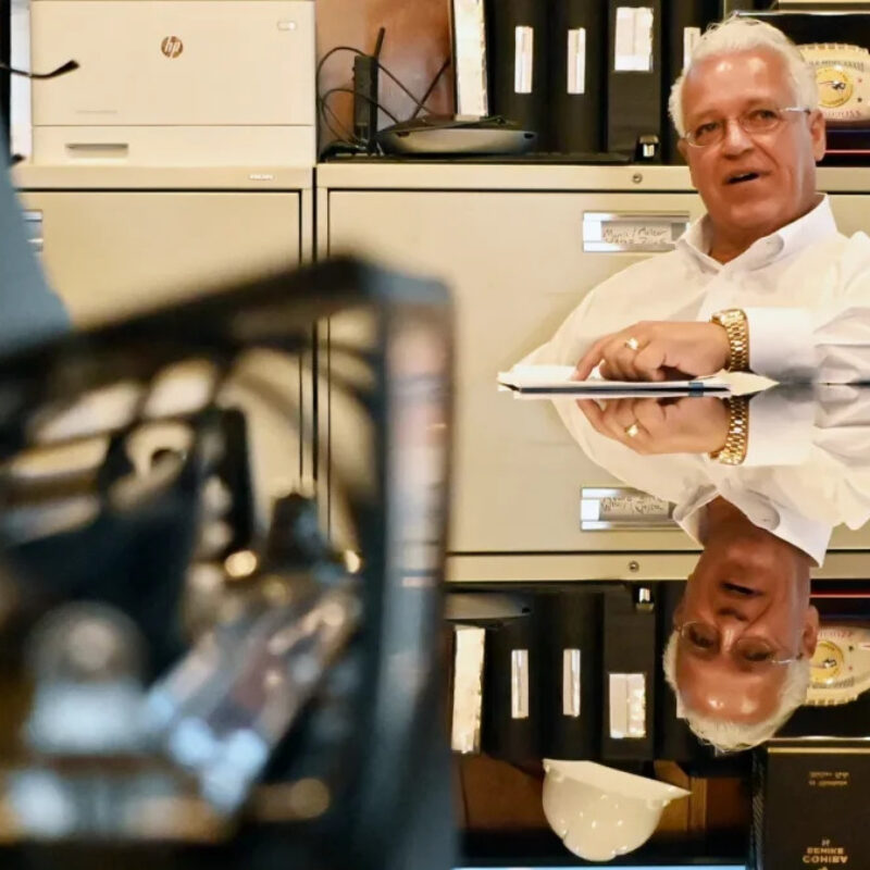 A man in a white shirt sits at a desk, holding a notepad, with office supplies and cabinets in the background.