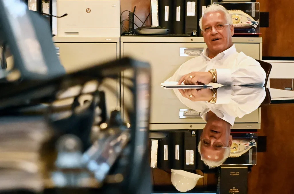 A man in a white shirt sits at a desk, holding a notepad, with office supplies and cabinets in the background.
