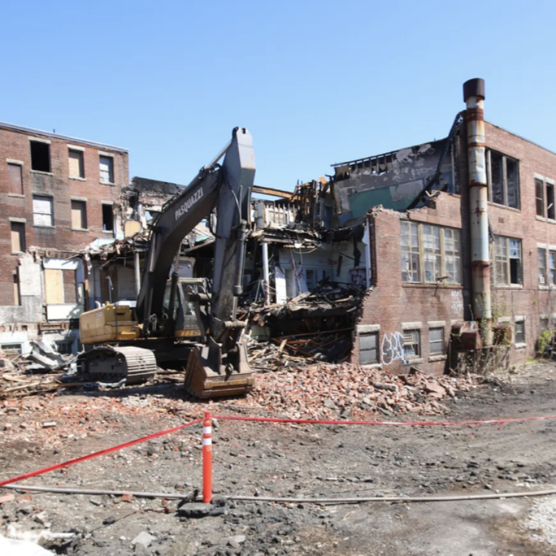 Excavator demolishes a partially collapsed brick building, surrounded by debris and construction barriers.