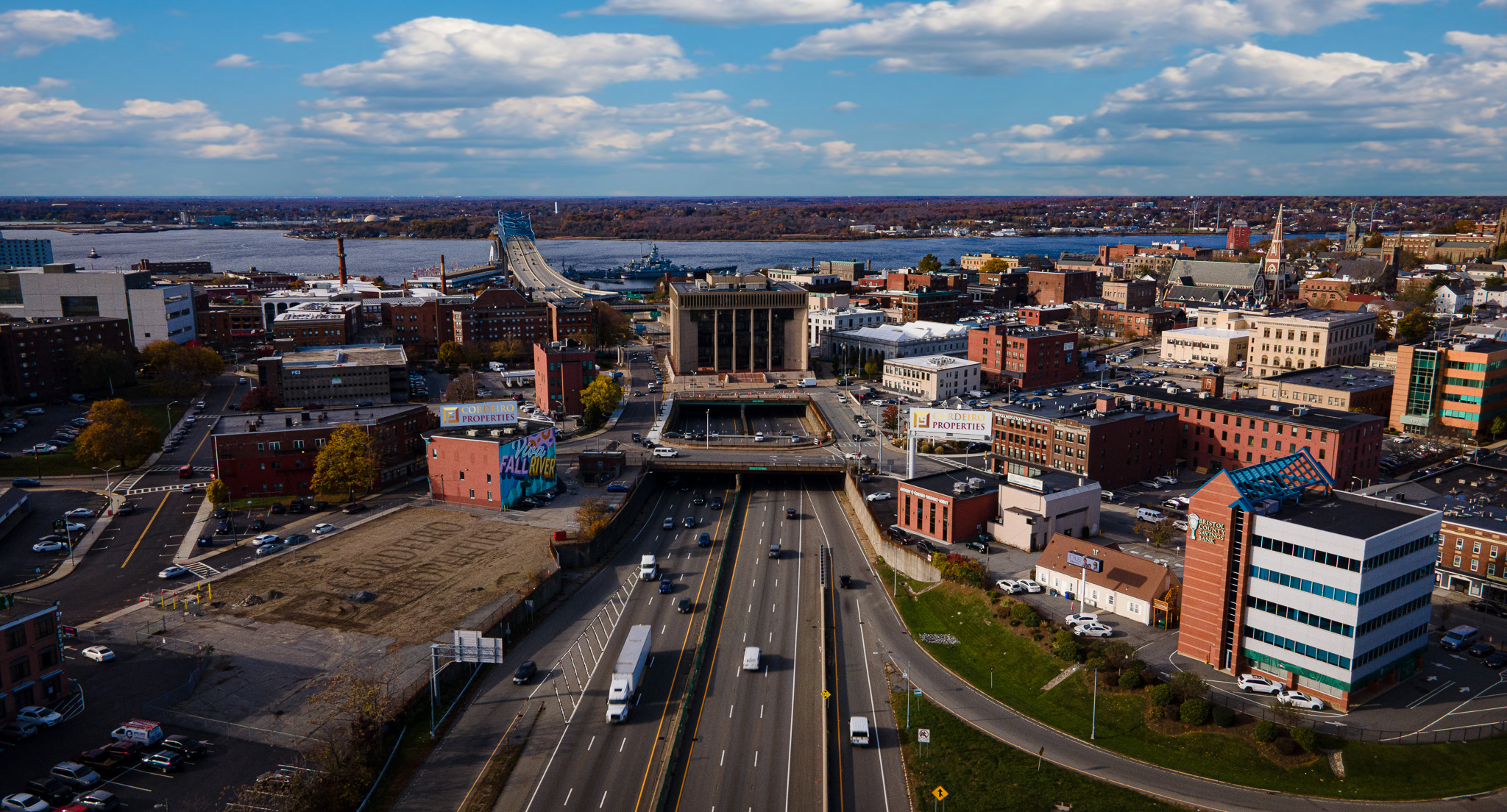 Aerial view of a city with buildings, a highway, and a river under a partly cloudy sky.