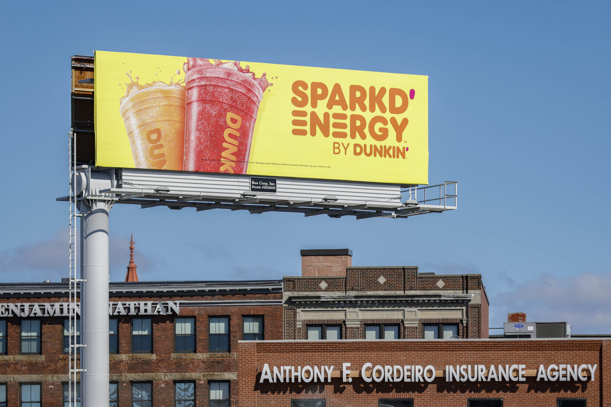 Billboard displays two colorful drinks with "SPARKD' ENERGY by DUNKIN'" text above a brick building.