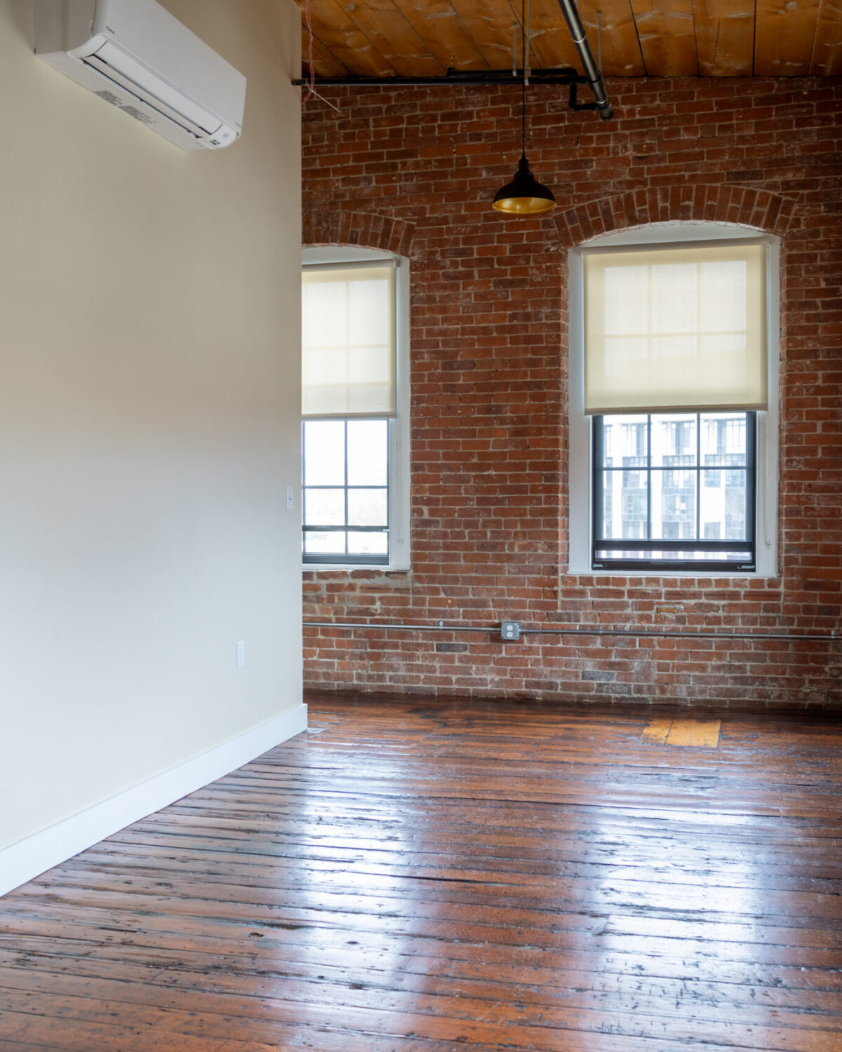 Empty room with exposed brick walls, wooden floor, and two windows with blinds. An air conditioning unit is mounted on the wall.