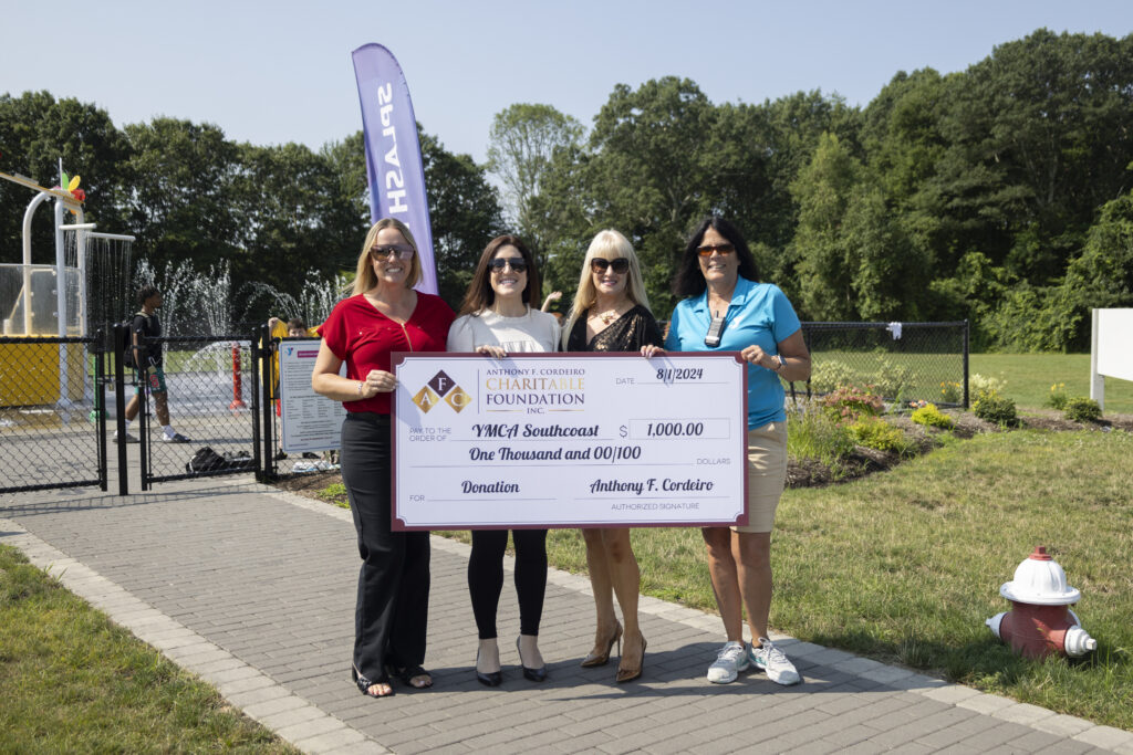 Four women hold a large donation check for $1,000 to YMCA Southcoast, with a splash pad in the background.
