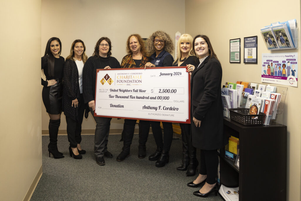 Seven women pose together, holding a large check for $2,500 to United Neighbors Fall River.