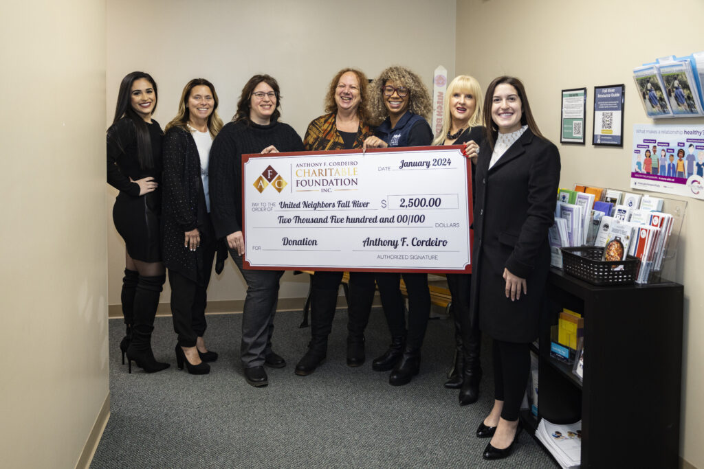 Seven women pose together, holding a large donation check for $2,500 to United Neighbors Fall River.