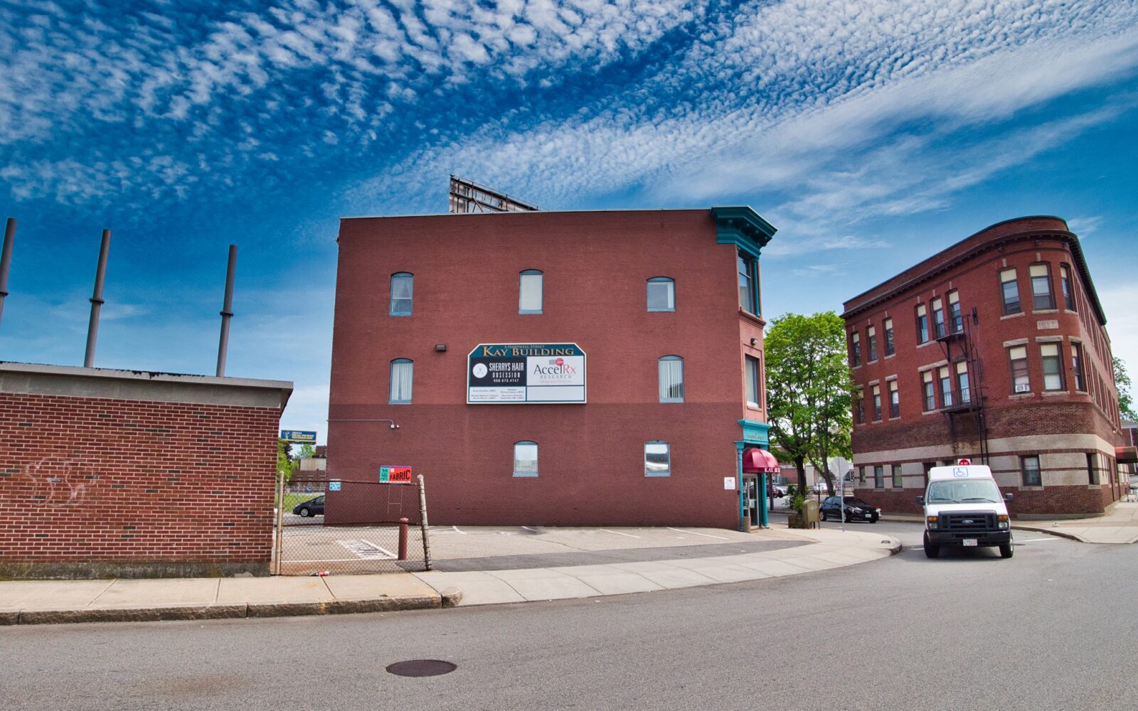 Red brick Kay Building with signage, adjacent to another building and a parked van on a clear day.