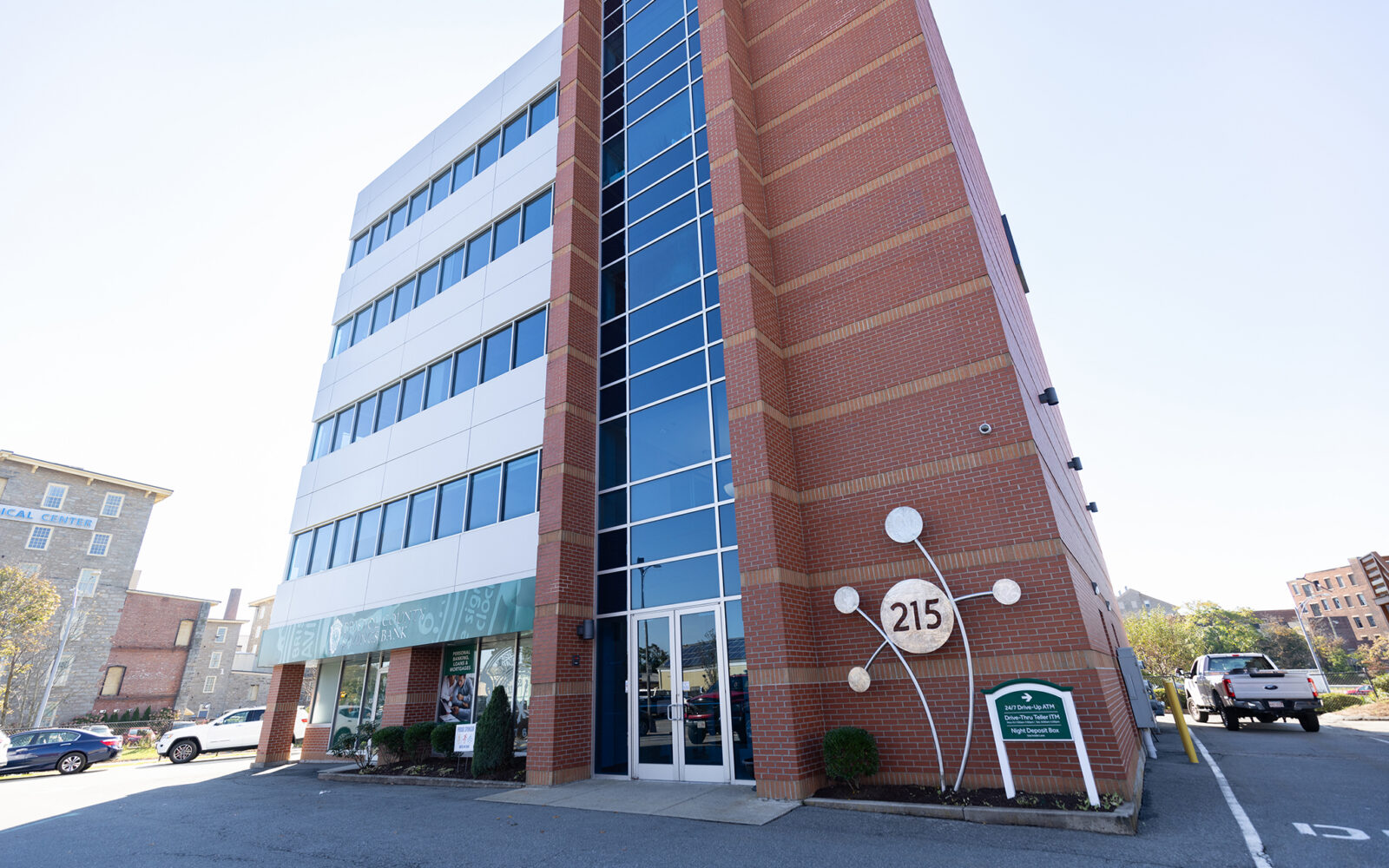 Five-story brick building with large glass entrance, featuring a sign displaying "215" and nearby parking area.