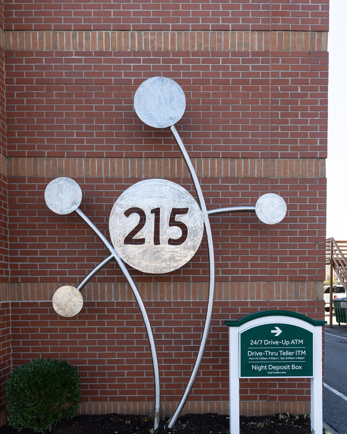 Metallic art installation featuring the number "215" on a brick wall, with a sign for bank services nearby.