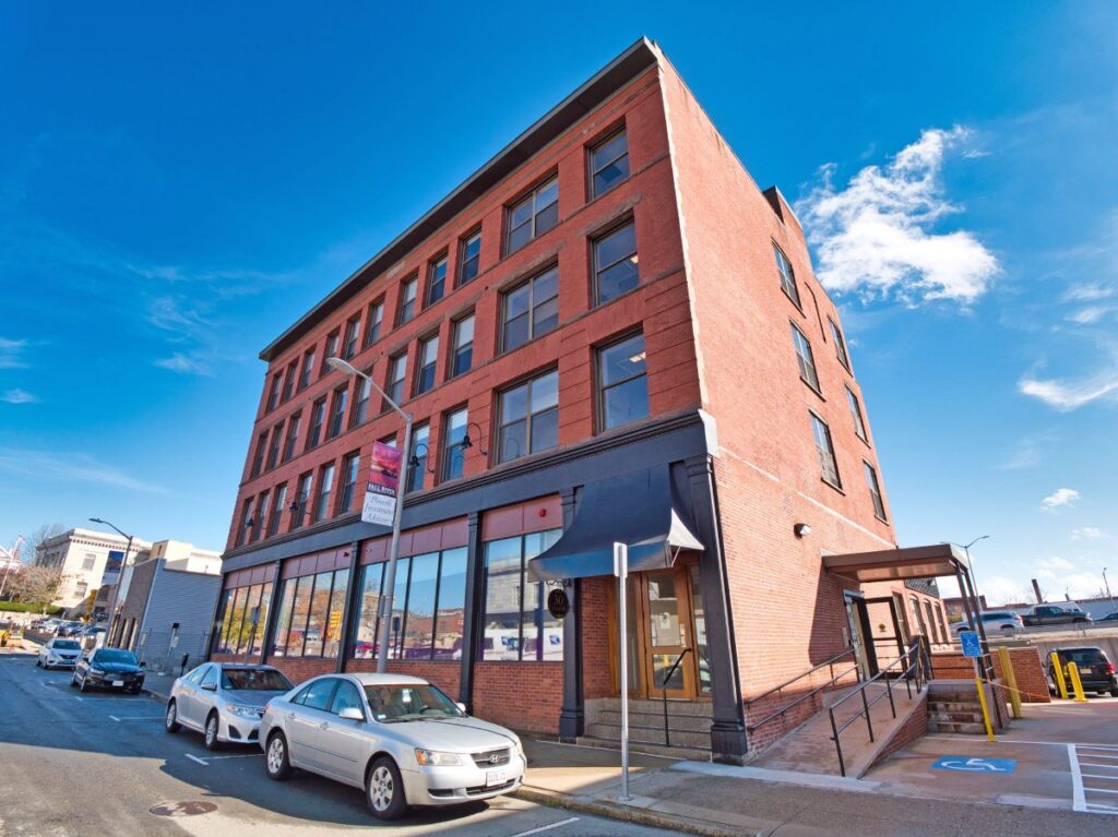 Red brick building with large windows, accessible entrance, and parked cars on a sunny street.