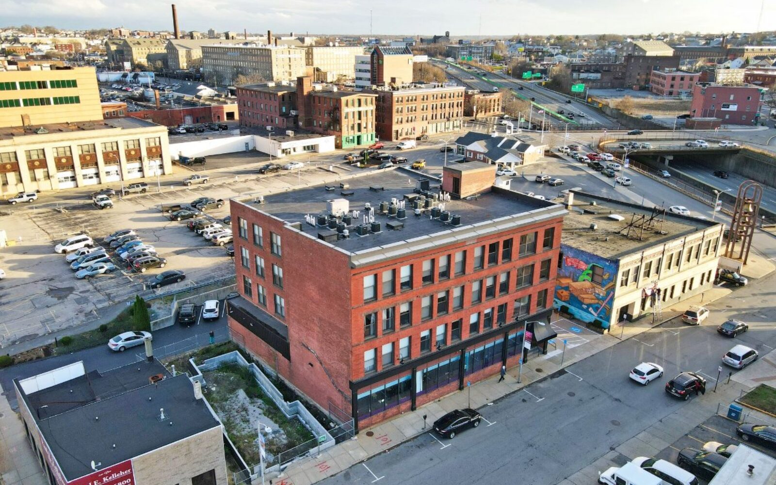 Aerial view of urban buildings, parking lots, and streets with vehicles and a colorful mural on a nearby structure.