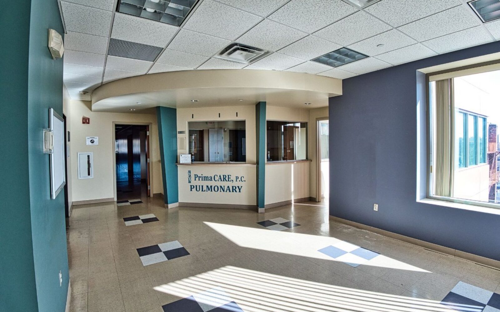 Bright, spacious waiting area with a reception desk labeled "PrimaCARE, P.C. Pulmonary." Sunlight streams through windows.