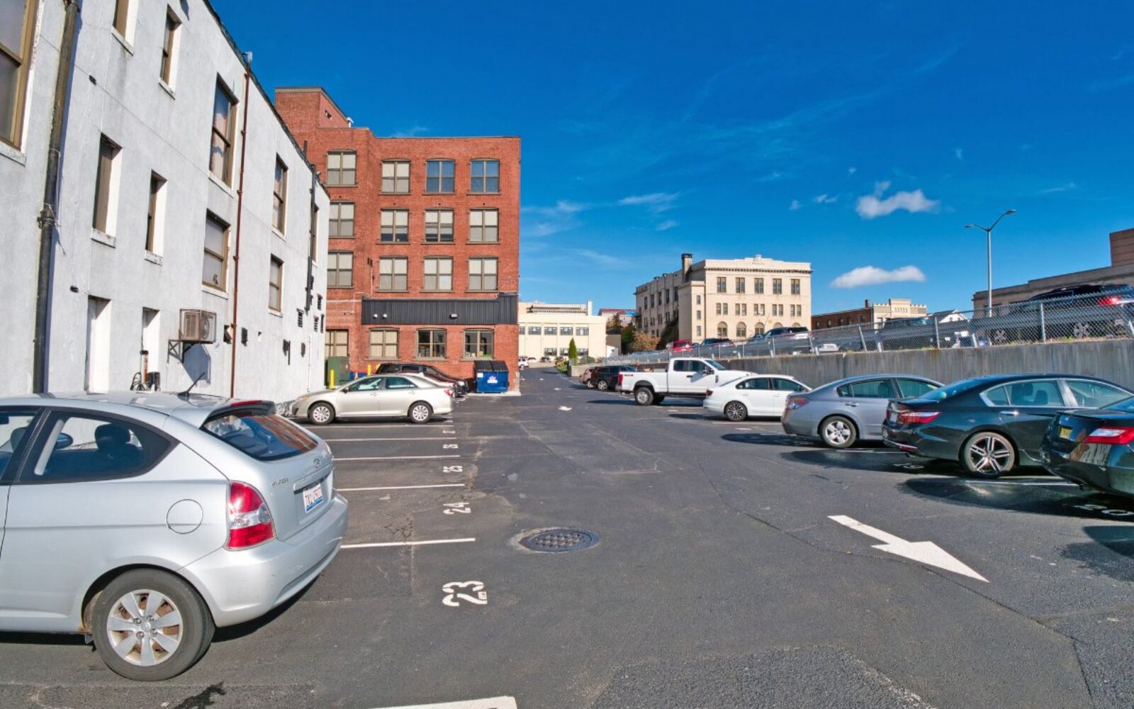 Parking lot with various cars, surrounded by brick and white buildings under a clear blue sky.