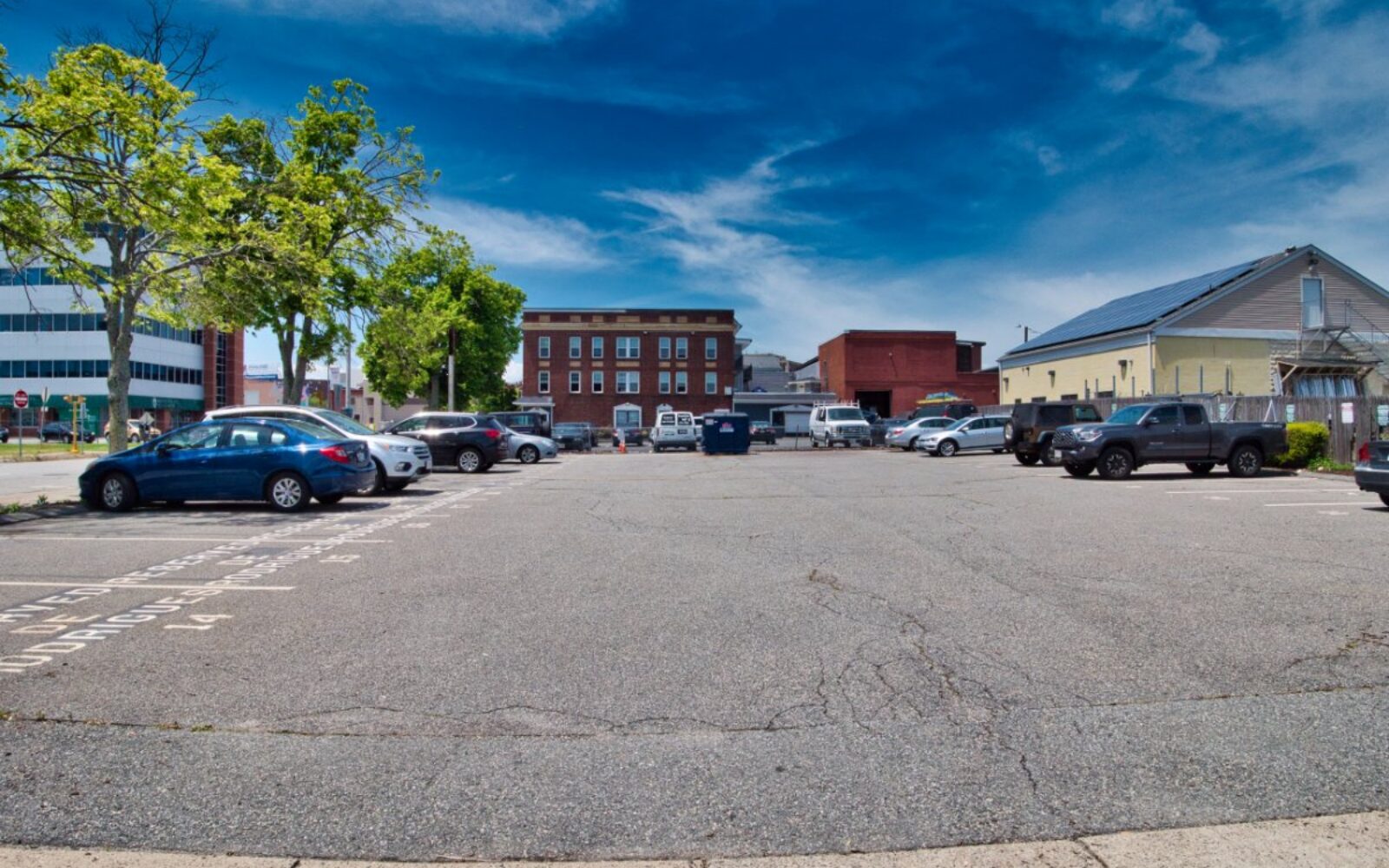 A parking lot with several cars, trees, and buildings under a clear blue sky.