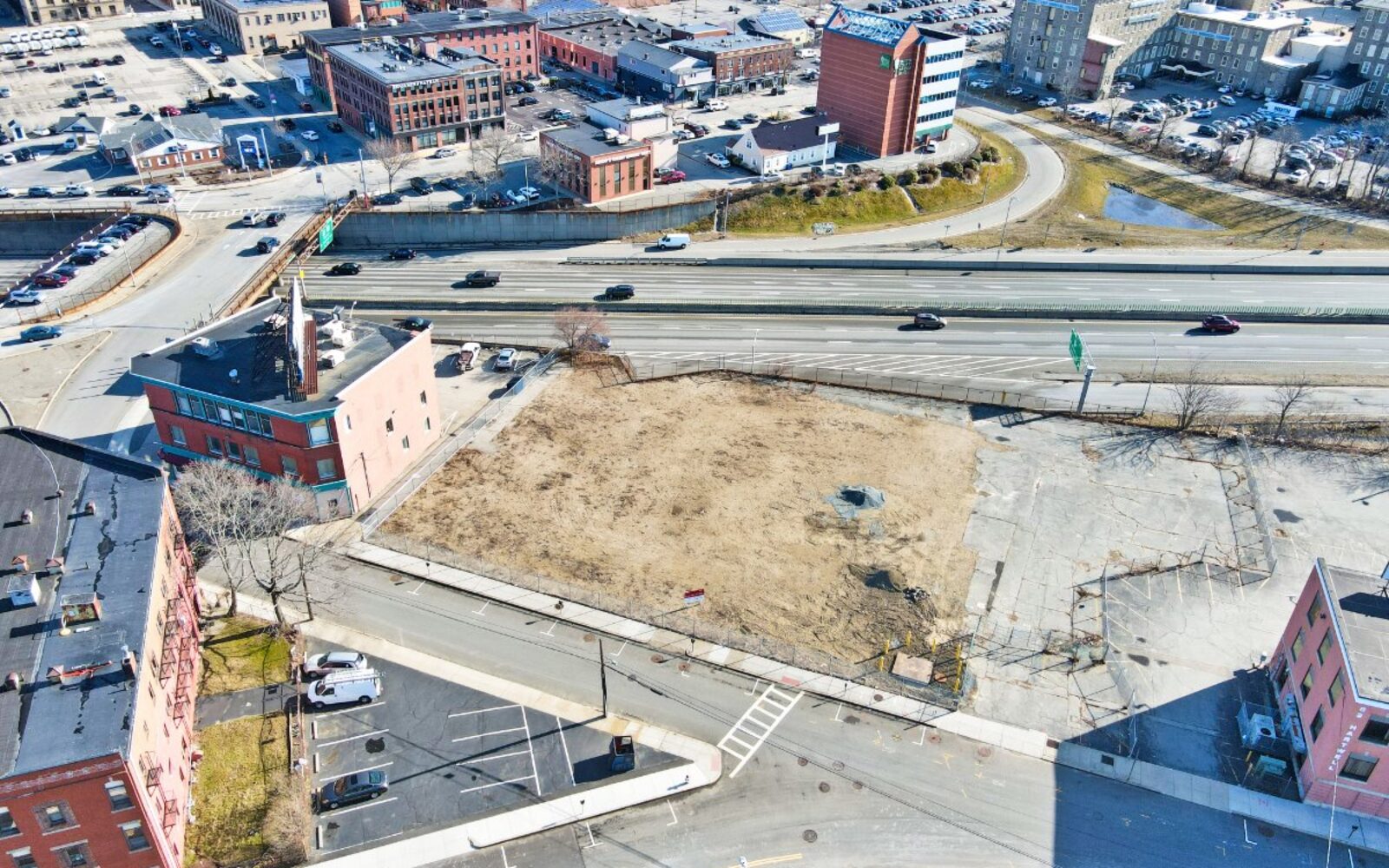 Aerial view of a vacant lot near a highway, surrounded by buildings and parked cars.