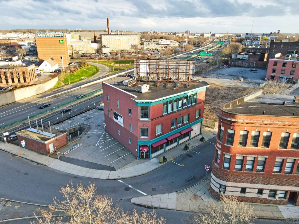 Aerial view of a red brick building with a green roof, surrounded by roads and vacant lots.