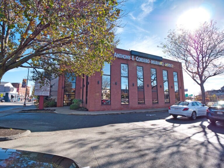 Brick building with large windows, featuring "Anthony F. Cordeiro Insurance Agency" sign. Trees and parked cars nearby.