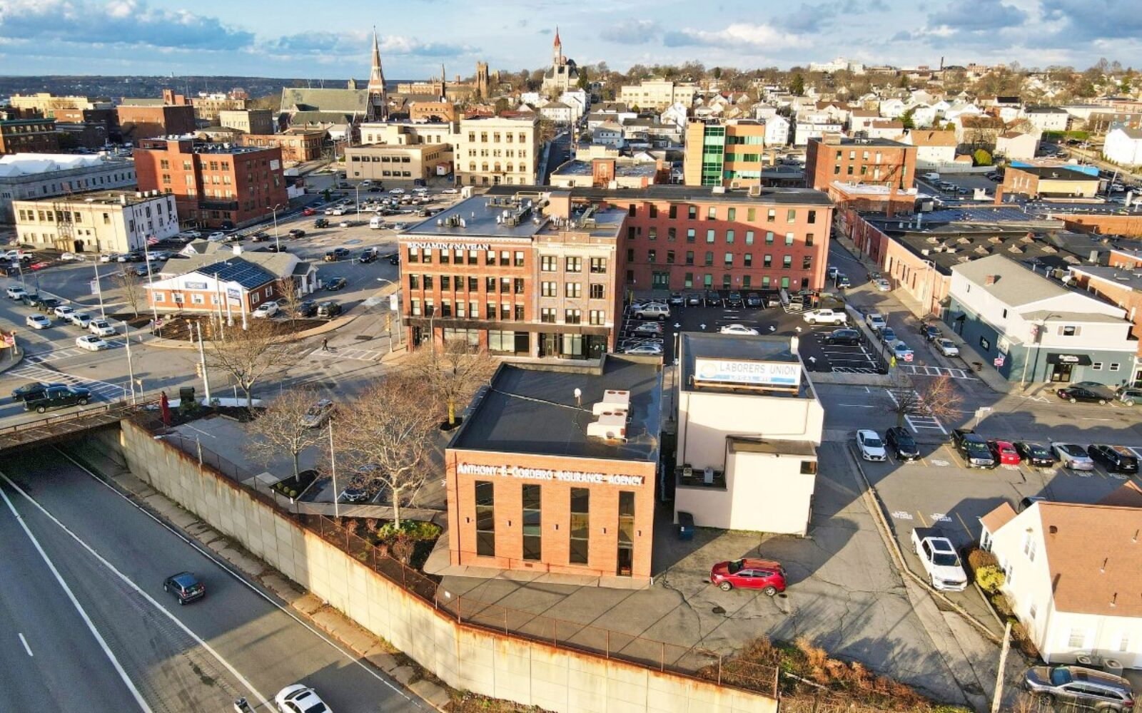 Aerial view of a city with various buildings, parking lots, and a highway at the bottom.