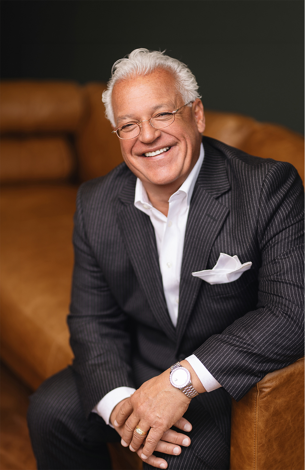 A smiling man in a pinstripe suit sits on a brown leather couch, resting his hands on his knees.