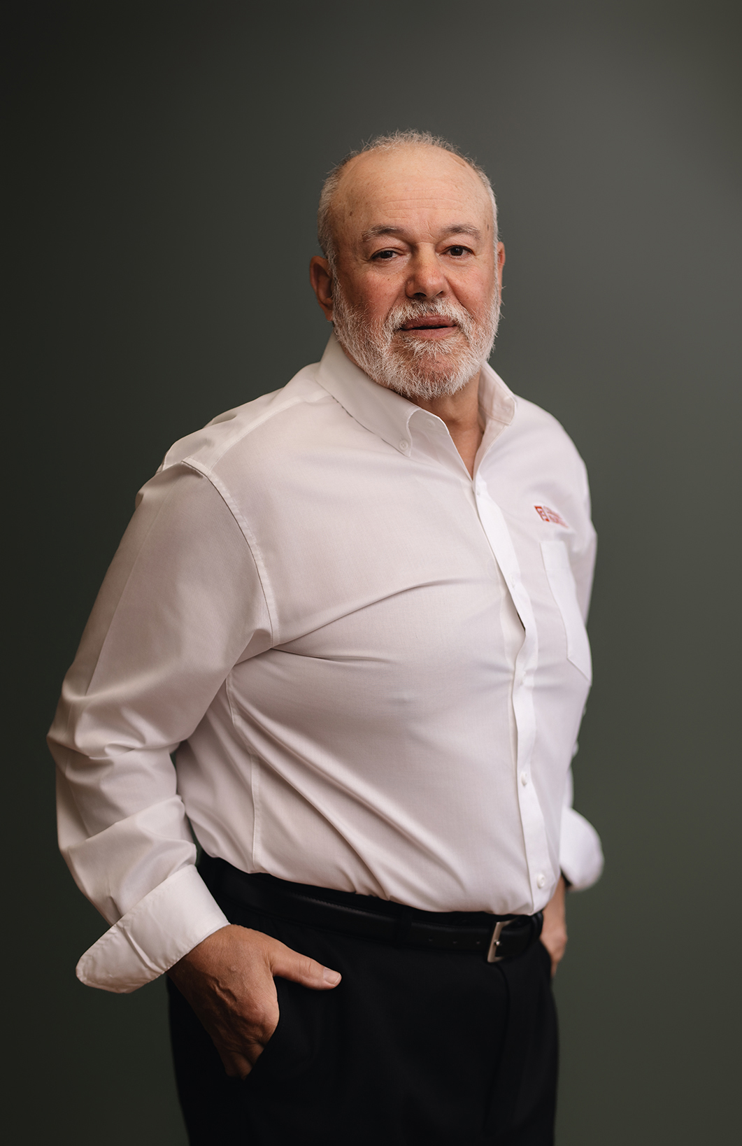 A man with a beard stands confidently, wearing a white shirt and black pants, against a dark green background.