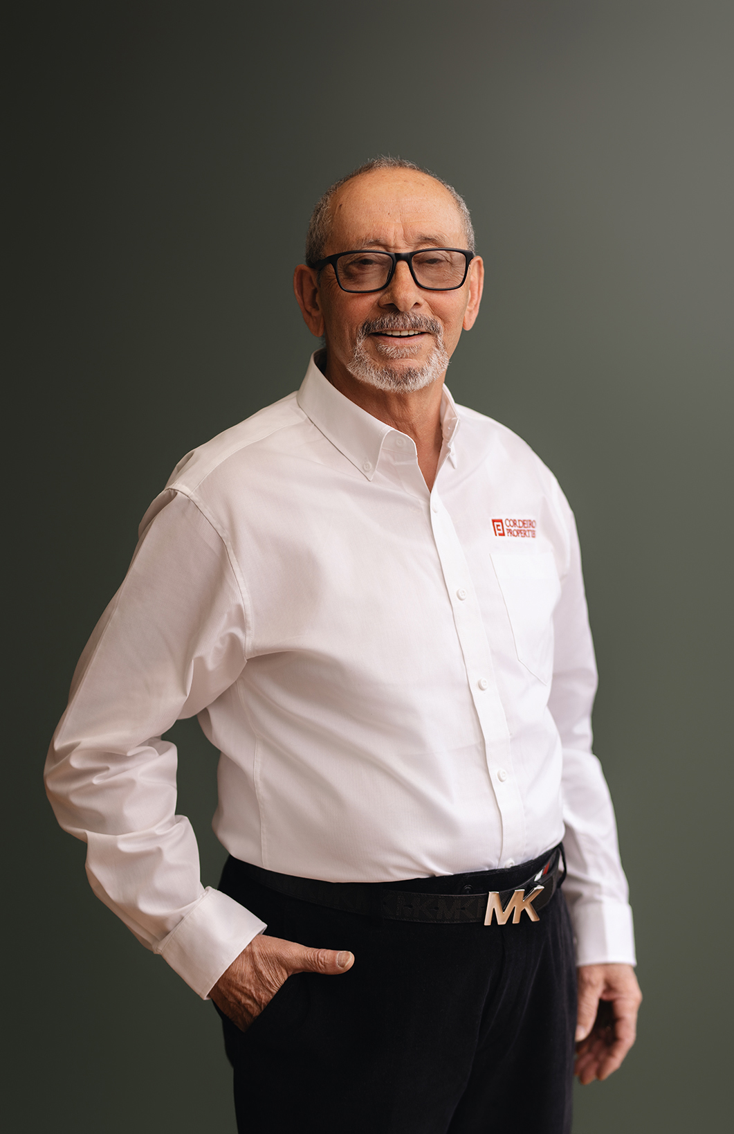 Older man in a white shirt and black pants, standing with one hand in his pocket, smiling at the camera.