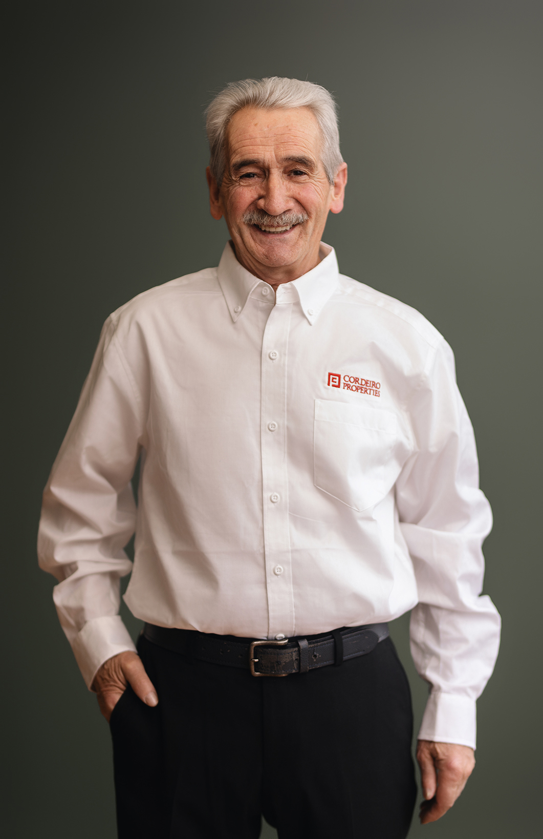A smiling older man in a white shirt with a logo, standing with hands in pockets against a gray background.