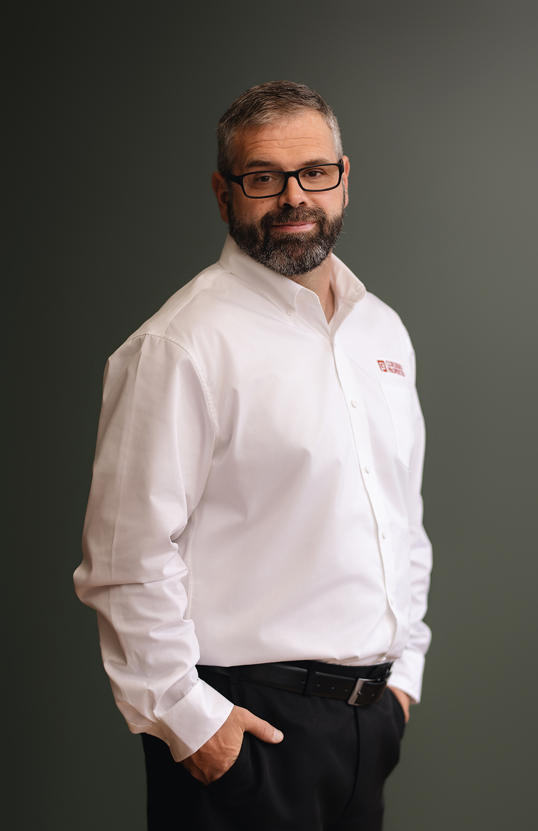 A man with glasses and a beard stands confidently, wearing a white shirt and black pants against a dark background.