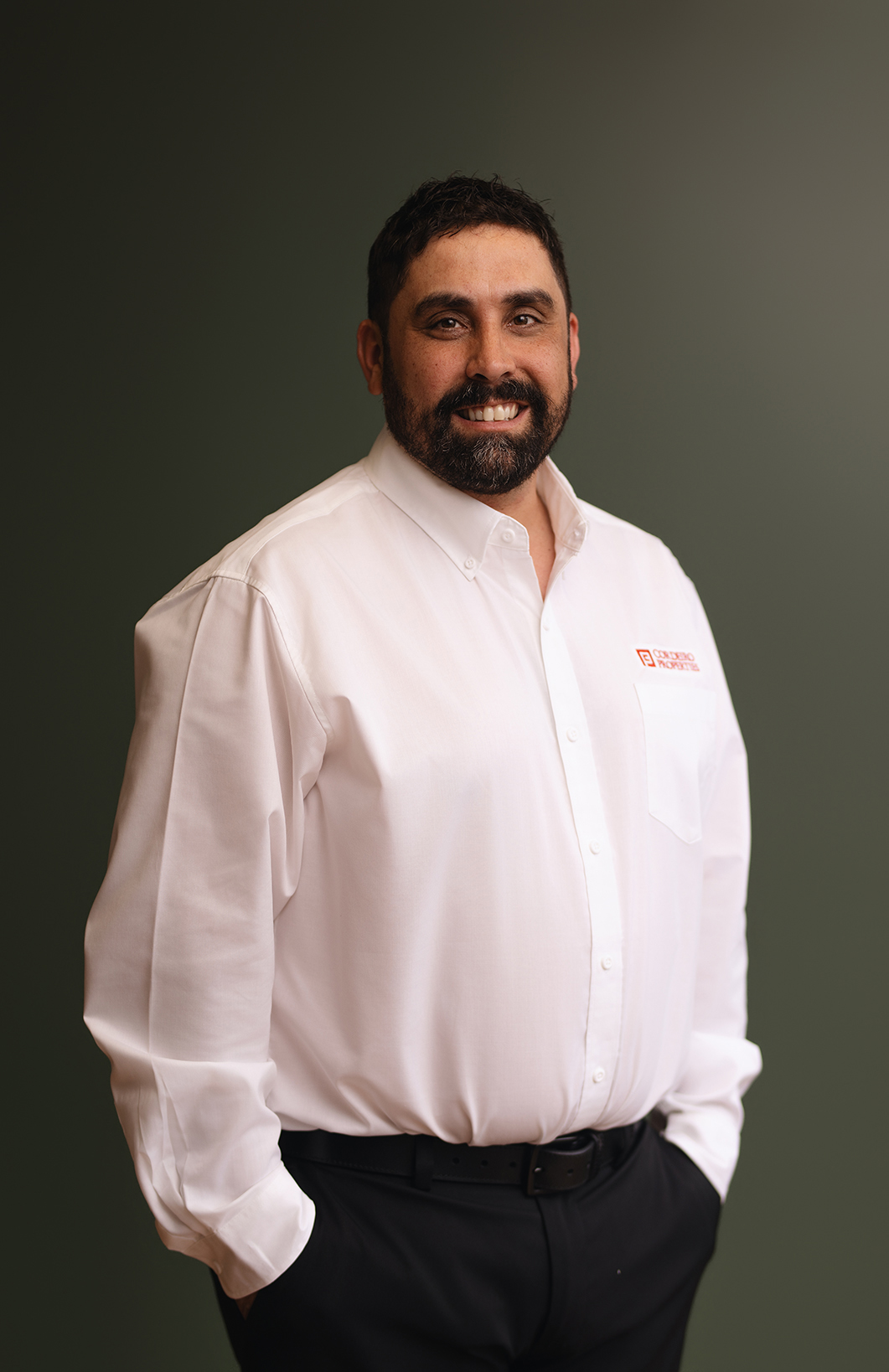 A man with a beard smiles, wearing a white shirt and black pants, standing against a green background.