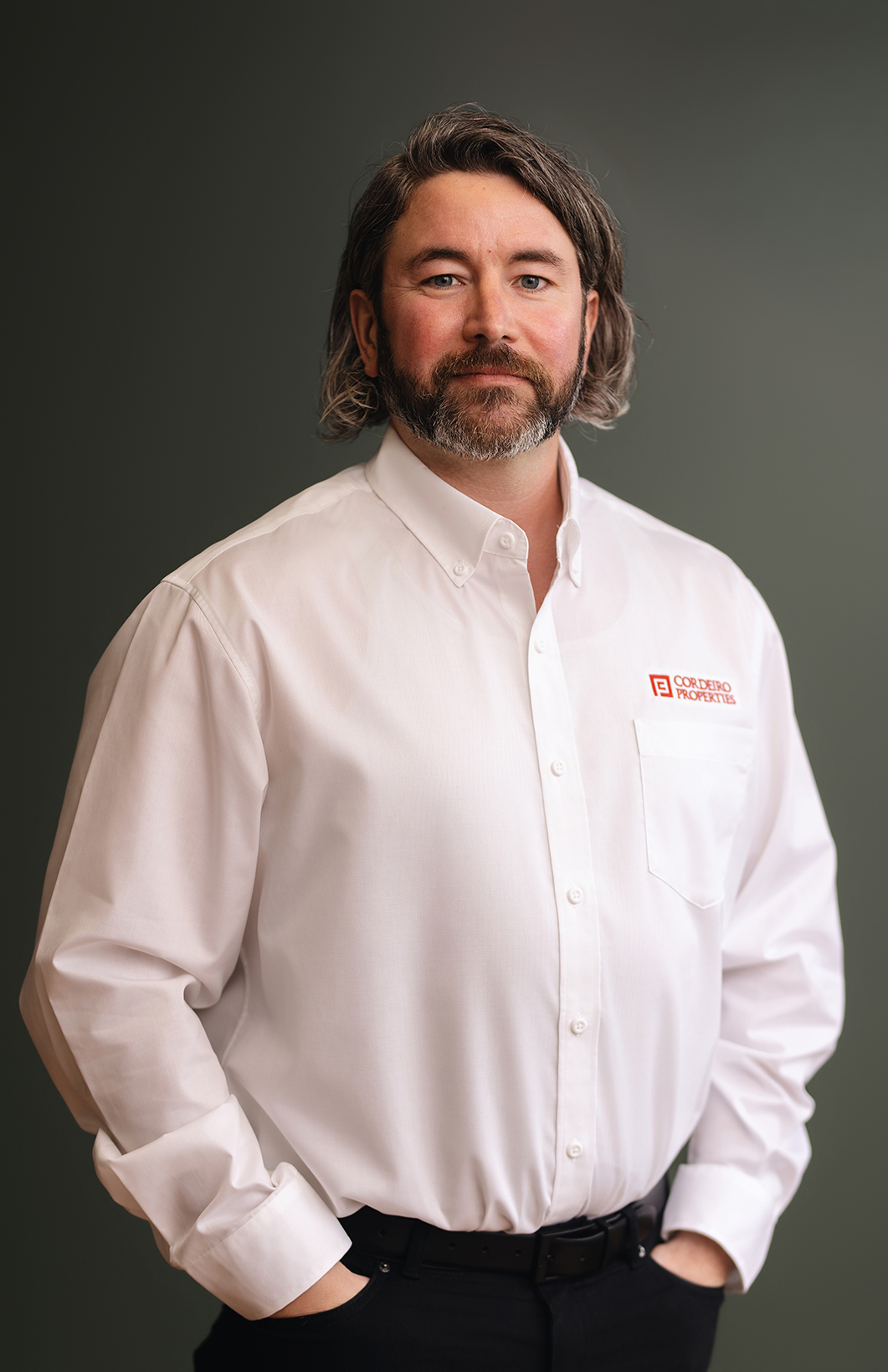 A man with long hair and a beard stands confidently, wearing a white shirt with a logo.