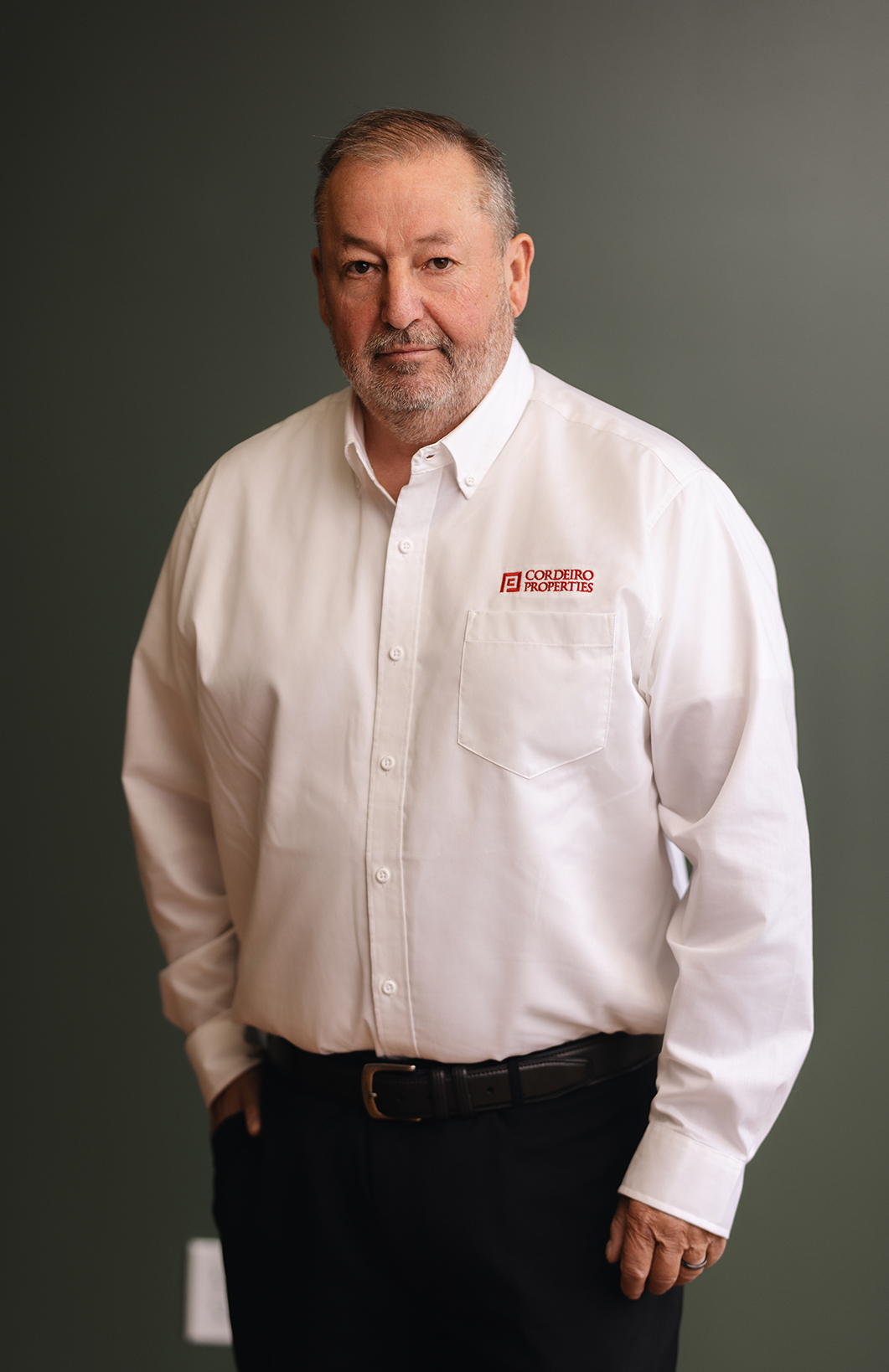 A man with a beard stands confidently, wearing a white shirt with a logo, against a green background.
