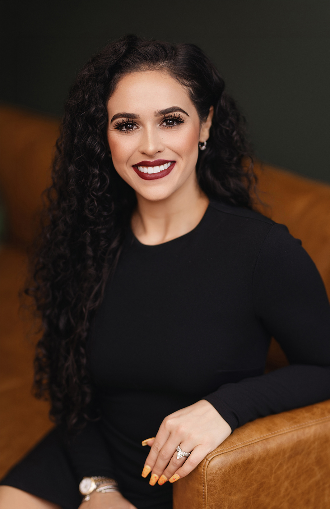 A woman with curly hair smiles while sitting on a brown couch, wearing a black dress and showcasing colorful nails.