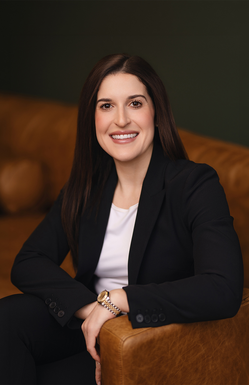 A woman in a black blazer smiles while sitting on a brown leather couch.
