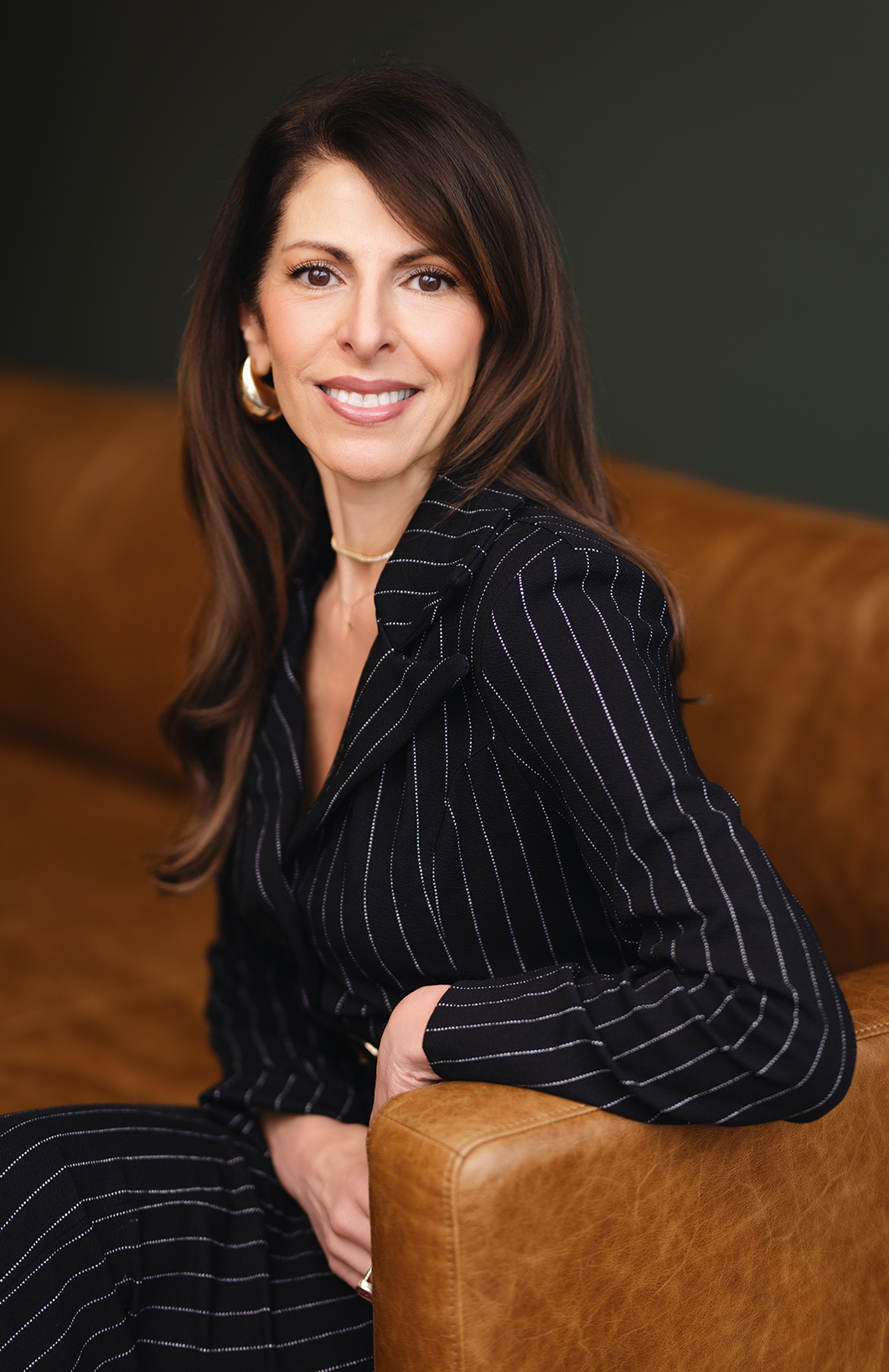 A woman with long hair smiles while seated on a brown leather couch, wearing a pinstriped suit.
