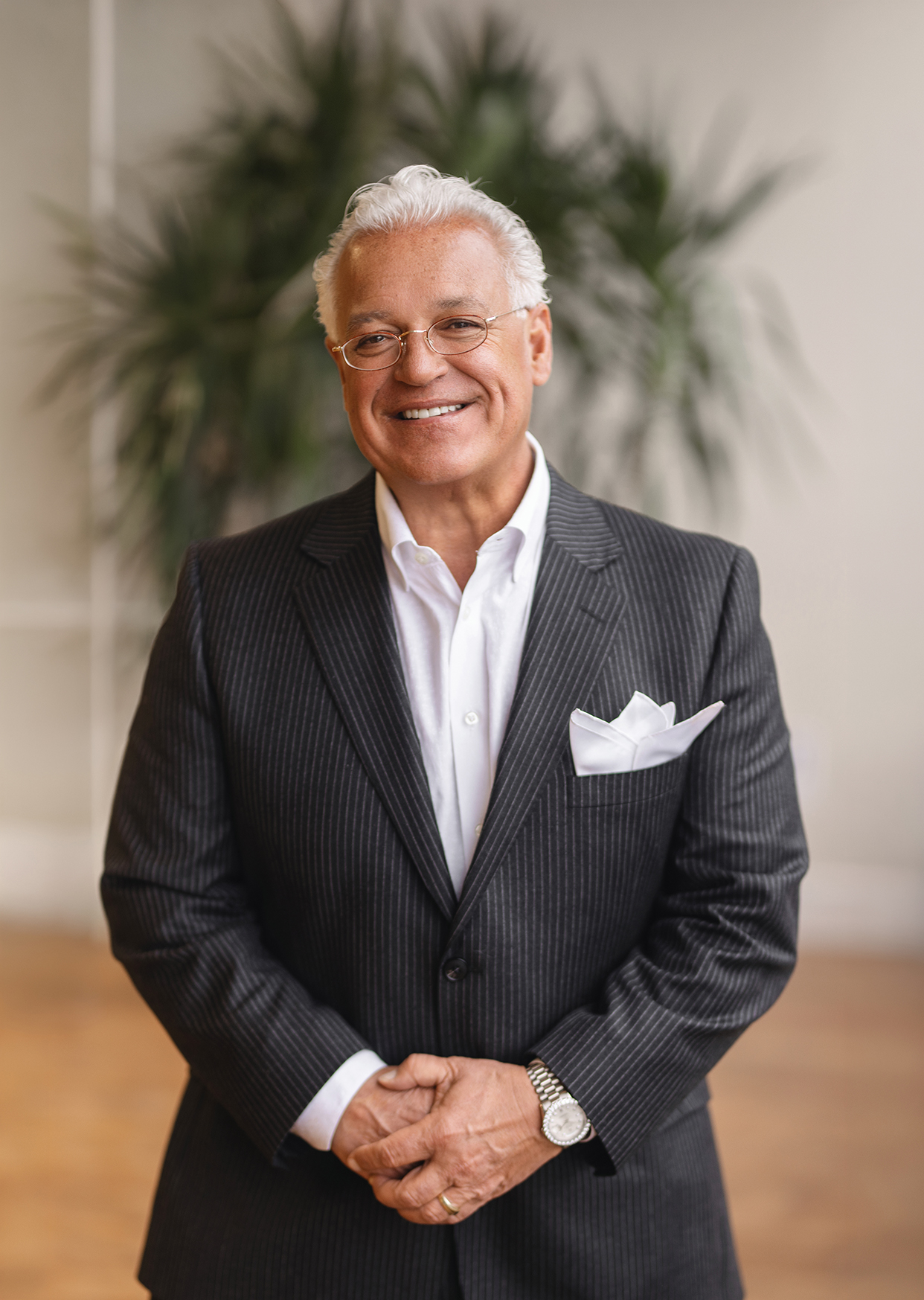 Smiling man in a pinstriped suit stands with hands clasped, a plant in the background.