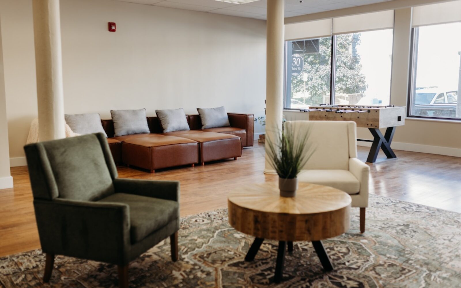 A cozy lounge area with two armchairs, a coffee table, and a foosball table near large windows.