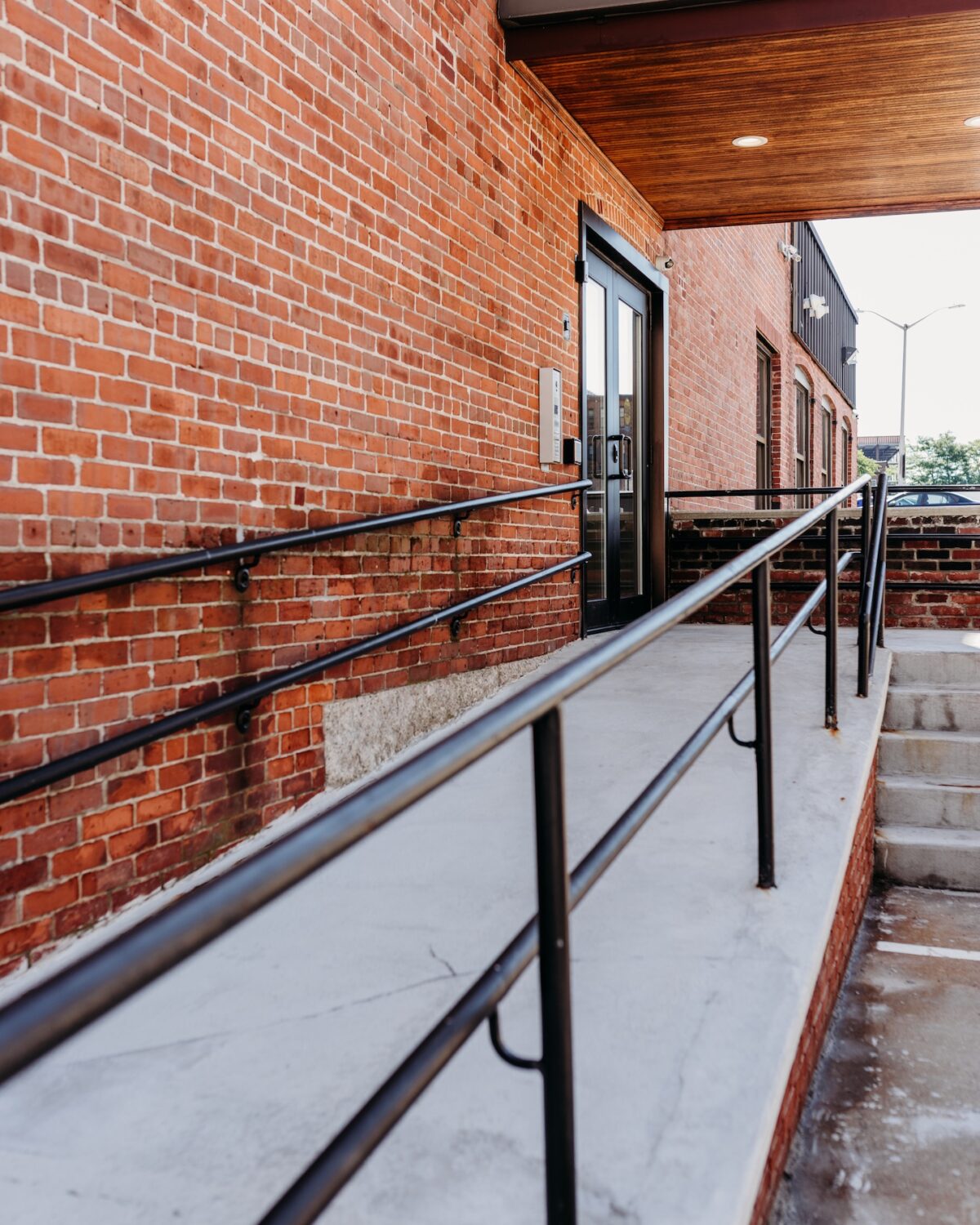 A sloped walkway with handrails leads to a glass door of a brick building.