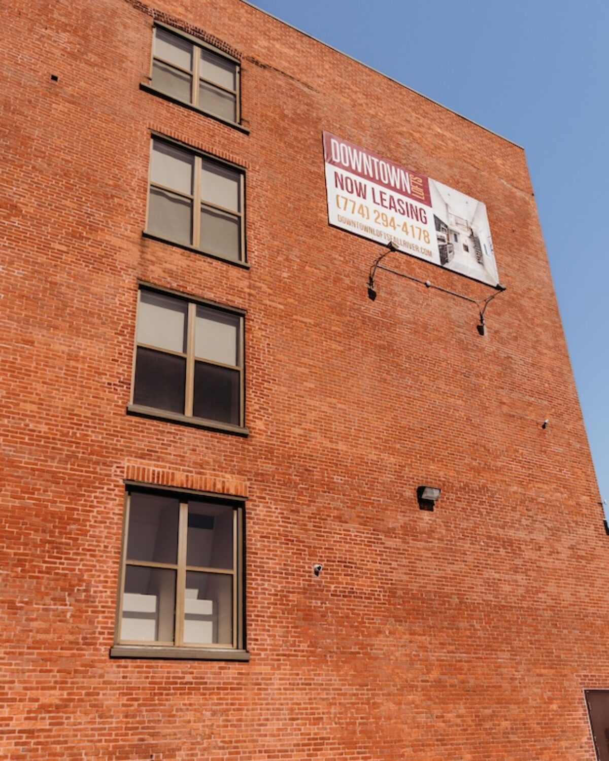 Red brick building with multiple windows and a leasing sign on the upper wall. Clear blue sky above.
