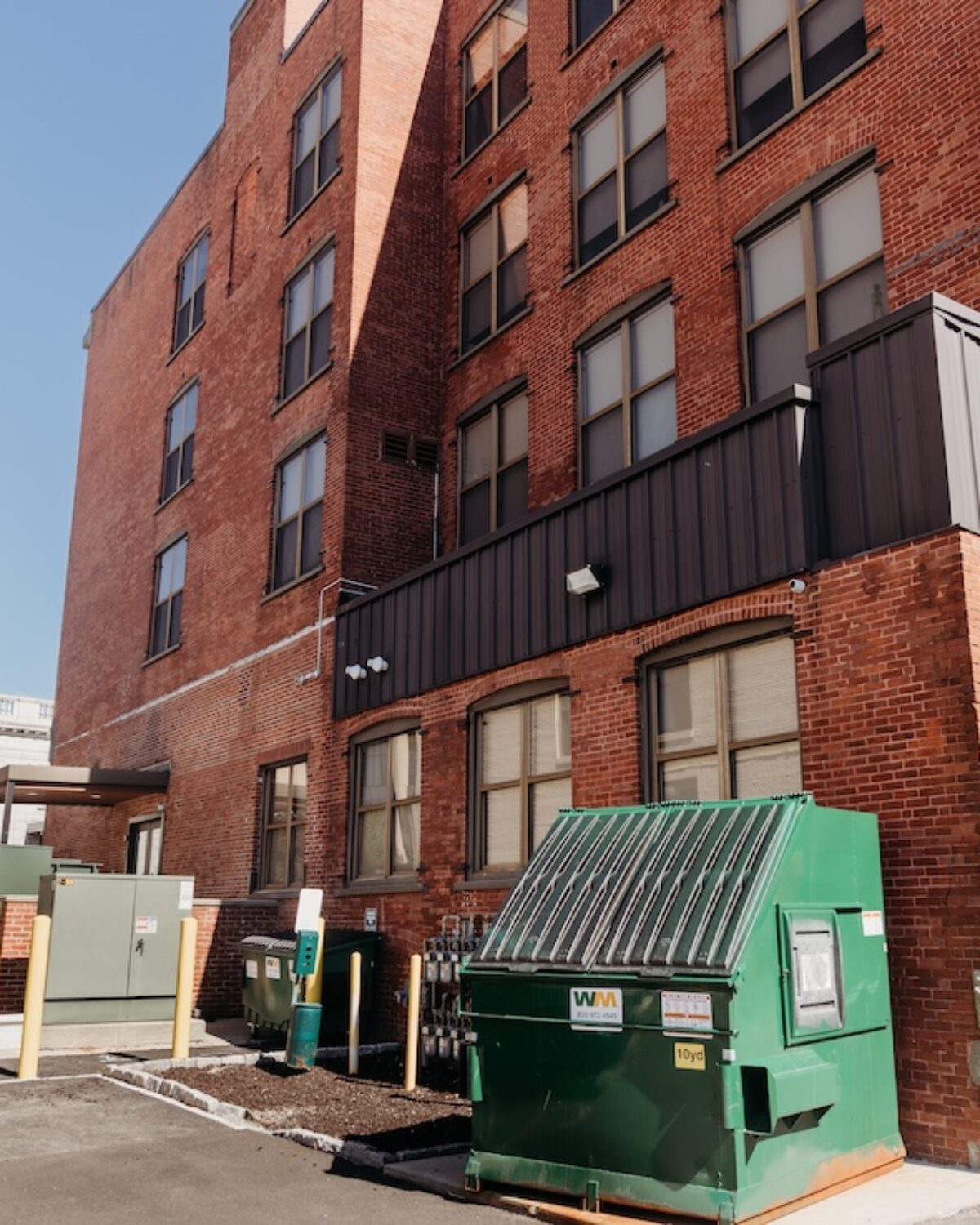 Brick building exterior with windows, green dumpster, and utility boxes in the parking area.