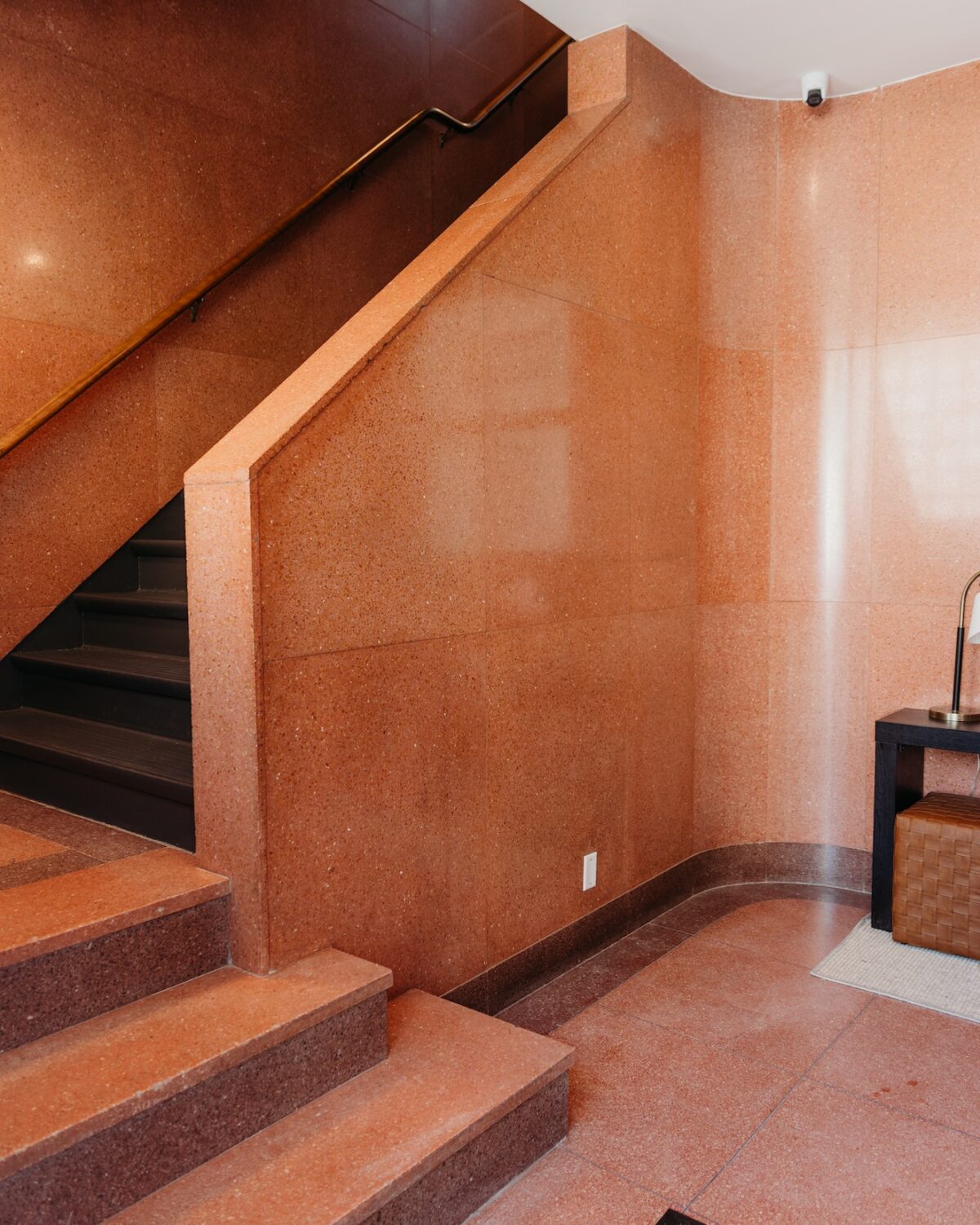 Interior view of a staircase with a wooden handrail, surrounded by polished reddish walls and a small table.